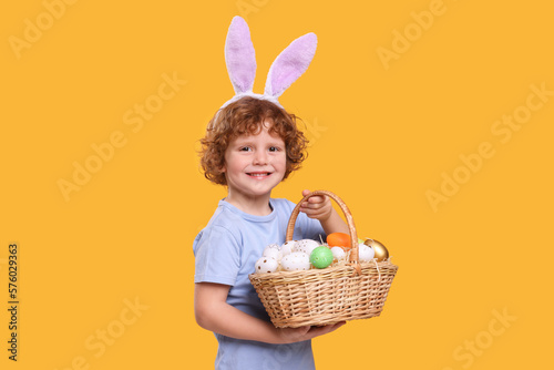 Portrait of happy boy in cute bunny ears headband holding wicker basket with Easter eggs on orange background