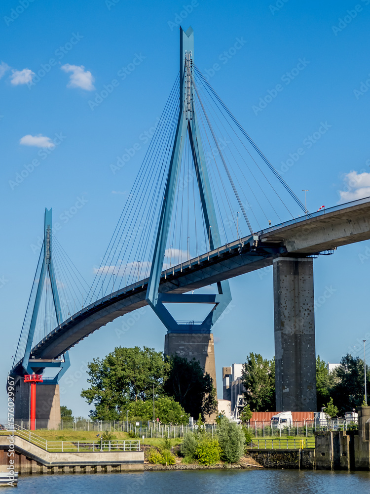 Obraz premium Portrait view of the tall blue dilapidated Köhlbrandbrücke suspension bridge seen from beneath from the riverside of harbor Rugenberger Hafen, landmark of the Port of Hamburg at a sunny summer day.