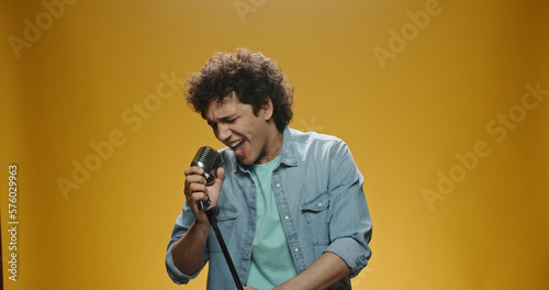 Young teenage Asian boy with curly hair in casual clothes singer performing at a concert. Man singing in microphone, isolated on yellow background 