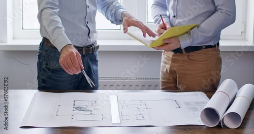 Two architects work next to a blueprint of a house, measuring and recording the values. Engineers discuss drawing a house on paper.