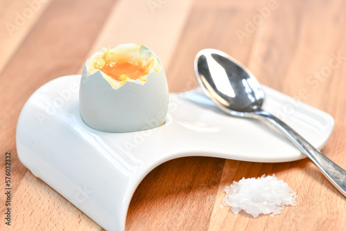 a single boiled egg in an egg cup broken open showing yolk with a spoon on a wooden surface