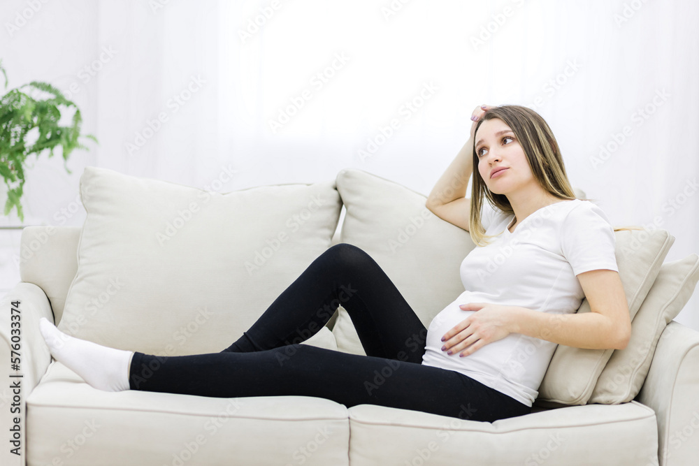 Young pregnant woman thinking about something on white sofa.