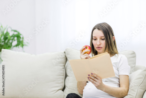 Photo of pregnant woman reading a book and eating an apple.