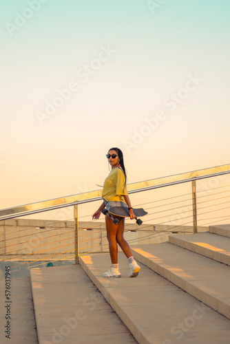 Pretty young woman with brown skin with long black braids standing holding a skateboard with her hand dressed in warm colored summer clothes on a concrete stairs on the seafront at summer sunset.
