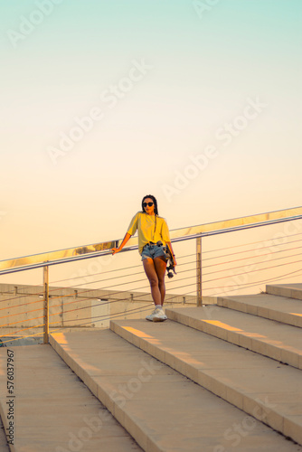 Pretty young woman with brown skin with long black braids standing holding a skateboard with her hand dressed in warm colored summer clothes on a concrete stairs on the seafront at summer sunset. © Mireia