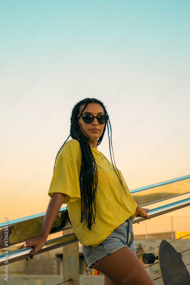 Pretty young woman with brown skin with long black braids standing with a skateboard by her side dressed in warm colored summer clothes on a concrete stairs on the seafront at summer sunset.