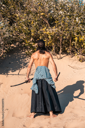 A Japanese warrior in a traditional kimano armed with a katana sword on a sandy shore next to the ocean during sunset. A man from medieval Asia. Reconstruction of cultural heritage. Culture in Japan. 