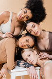 young multicultural women in lingerie posing near stool and looking at camera isolated on beige.