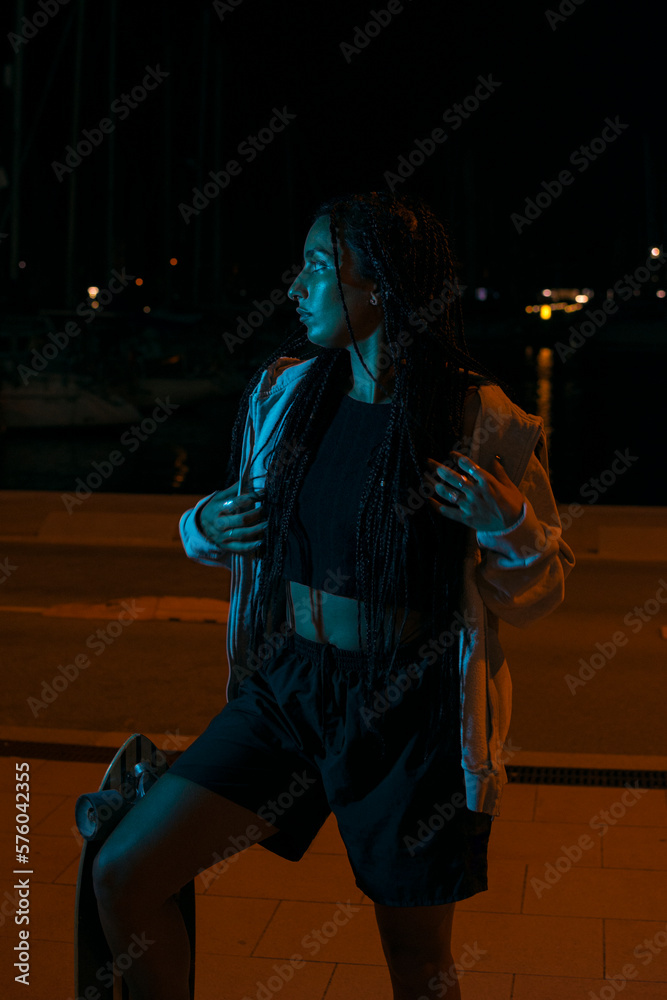 Naklejka premium Pretty young woman with brown skin with long black braids holding her skateboard dressed in a gray sweatshirt and dark clothes posing on the seafront on a summer night with blue led light.