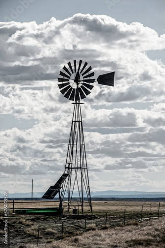 Windmill on the plains, powered by solar panels with livestock watering tank by barbed wire fence with mountains on horizon- vintage desaturated