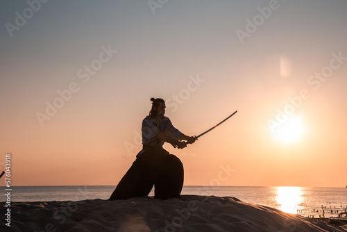 A Japanese warrior in a traditional kimano armed with a katana sword on a sandy shore next to the ocean during sunset. A man from medieval Asia. Reconstruction of cultural heritage. Culture in Japan.  photo