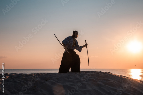 A Japanese warrior in a traditional kimano armed with a katana sword on a sandy shore next to the ocean during sunset. A man from medieval Asia. Reconstruction of cultural heritage. Culture in Japan. 