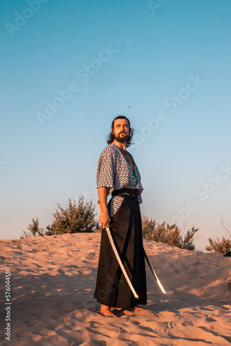 A Japanese warrior in a traditional kimano armed with a katana sword on a sandy shore next to the ocean during sunset. A man from medieval Asia. Reconstruction of cultural heritage. Culture in Japan. 