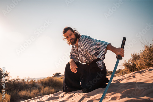 A Japanese warrior in a traditional kimano armed with a katana sword on a sandy shore next to the ocean during sunset. A man from medieval Asia. Reconstruction of cultural heritage. Culture in Japan. 