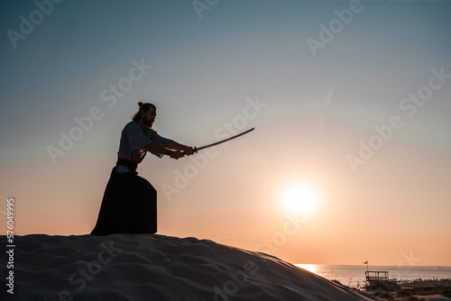 A Japanese warrior in a traditional kimano armed with a katana sword on a sandy shore next to the ocean during sunset. A man from medieval Asia. Reconstruction of cultural heritage. Culture in Japan.  photo