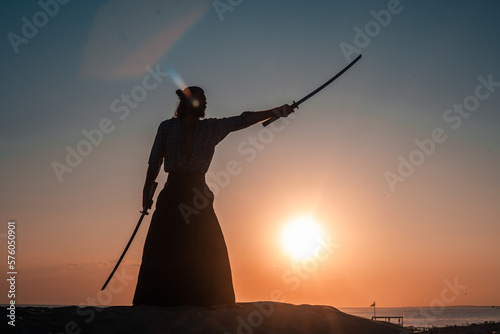 A Japanese warrior in a traditional kimano armed with a katana sword on a sandy shore next to the ocean during sunset. A man from medieval Asia. Reconstruction of cultural heritage. Culture in Japan. 