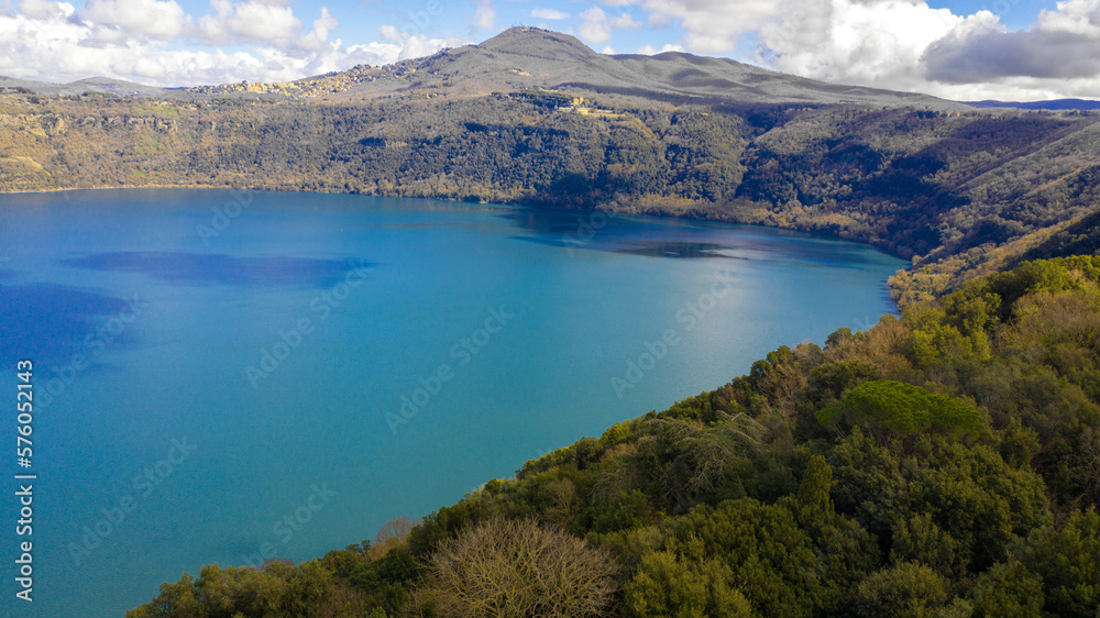 Aerial view of Lake Albano, a volcanic crater lake. A luxuriant forest grows around the water, which turns yellow and red in autumn.