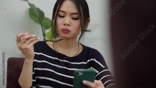 Young Smiling Woman Using Mobile Phone At Home While Eating a Soup. 