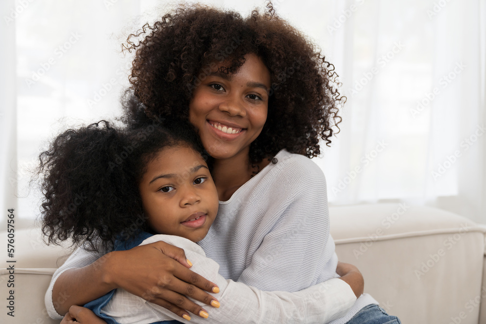 Happy afro family embracing together on sofa at home. Smiling African American young mother hugging her child daughter while sitting on couch at home Happy afro family embracing together on sofa at home. Smiling African American young mother hugging her child daughter while sitting on couch at home