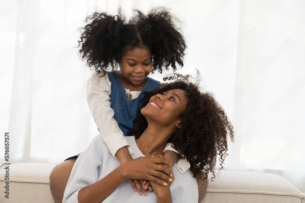 Obraz premium Happy afro family embracing together on sofa at home. Smiling African American young mother hugging her child daughter while sitting on couch at home