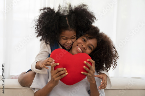 Happy afro family embracing together and surprise with pillow heart shape on sofa at home. Smiling African American young mother hugging daughter and spending time together while sitting on couch