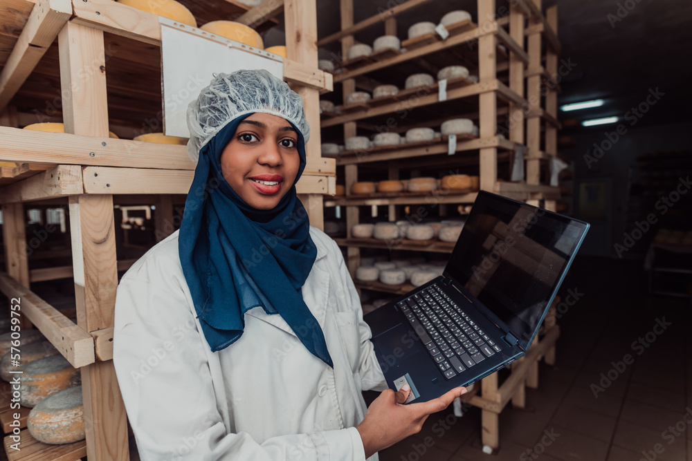 African American Muslim businesswoman checking product quality and entering data into a laptop at a local cheese manufacturing company