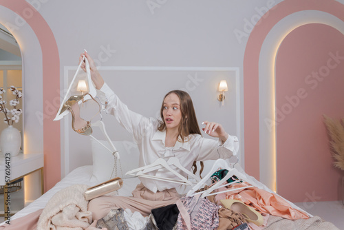 Young woman surrounded by different clothes and hangers in messy bedroom. many stacks of clothes. Disorder and mess. Clothing clutter and disorganization in the bedroom.