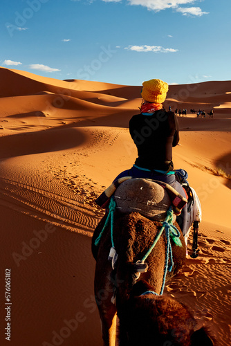 A young women in a yellow cap rides a camel  through the dunes in the Sahara Desert. View of the woman from behind, in the background, small silhouettes of other tourists. Merzouga, Morocco