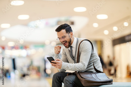 A young man is sitting on a bench in shopping mall and celebrating discount while smiling at the phone.
