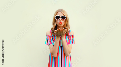 Summer portrait of beautiful young woman model blowing her lips sending sweet air kiss wearing heart shaped sunglasses on white background