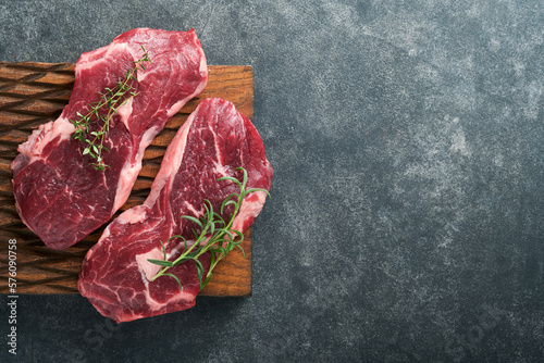 Raw beef steak. Marbled raw fresh Ribeye steak with rosemary, salt and pepper on wooden cutting board on dark concrete background. Raw beef steak and spices for cooking. Mock up.