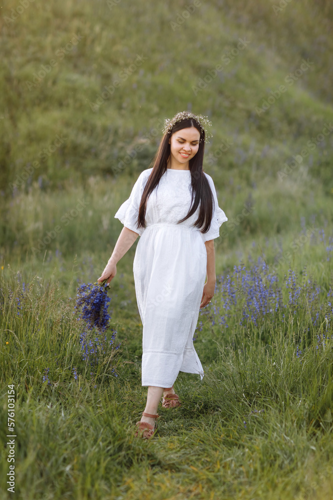 Young smiling beautiful woman in white dress and wreath of wildflowers with a bouquet of purple lupine flowers stands in green meadow in spring. Womens day. Summertime. Romantic mood. Nature concept