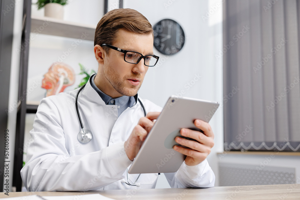 Male Caucasian doctor in white medical uniform filling patient medical history or anamnesis on tablet in office. Doctor consulting client online on tablet in modern clinic, healthcare concept - obrazy, fototapety, plakaty 
