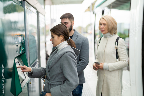 Young attractive woman using ticket vending machine at city tram stop in autumn day..