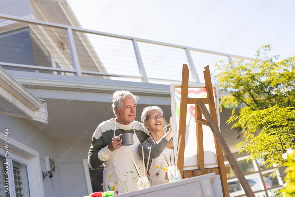 Happy senior diverse couple painting on easel in garden Happy senior diverse couple painting on easel in garden