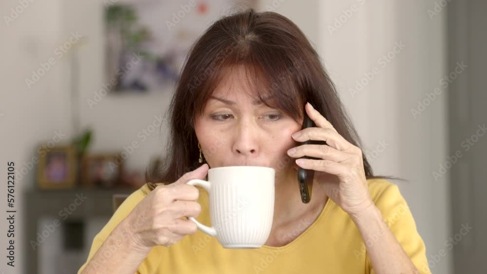 Frontal closeup beautiful mature woman long hair thin earrings in yellow top talking on mobile phone smiling drinking hot coffee winces face. Frontal closeup beautiful mature woman long hair thin earrings in yellow top talking on mobile phone smiling drinking hot coffee winces face.