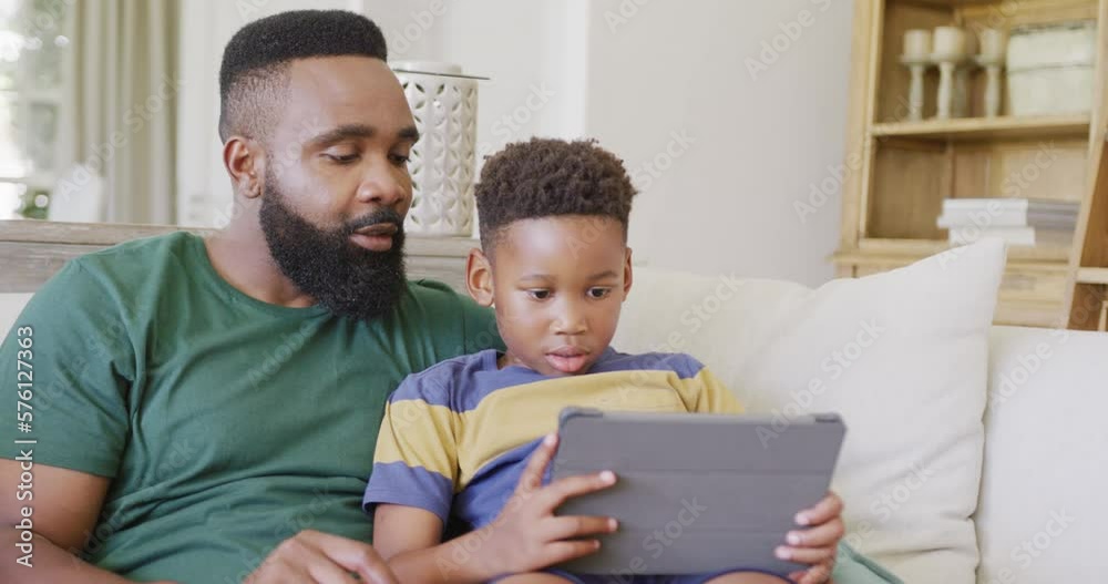 Happy african american father and son sitting on sofa, using tablet, in slow motion Happy african american father and son sitting on sofa, using tablet, in slow motion