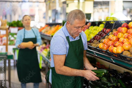 male seller of vegetable shop chooses fresh and ripe zucchini for regular customer