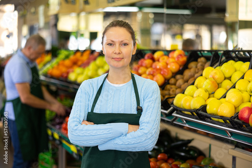 Joyful skilled adult female seller in uniform posing during working day in middle of grocery shop