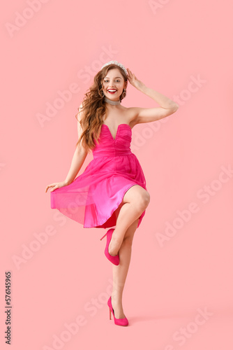 Young woman in tiara and bright prom dress dancing on pink background