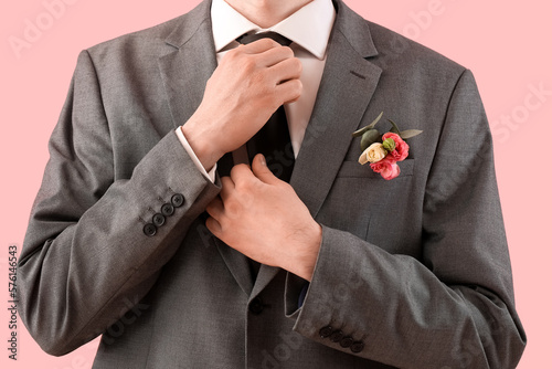 Young man in prom suit with boutonniere fixing necktie on pink background, closeup