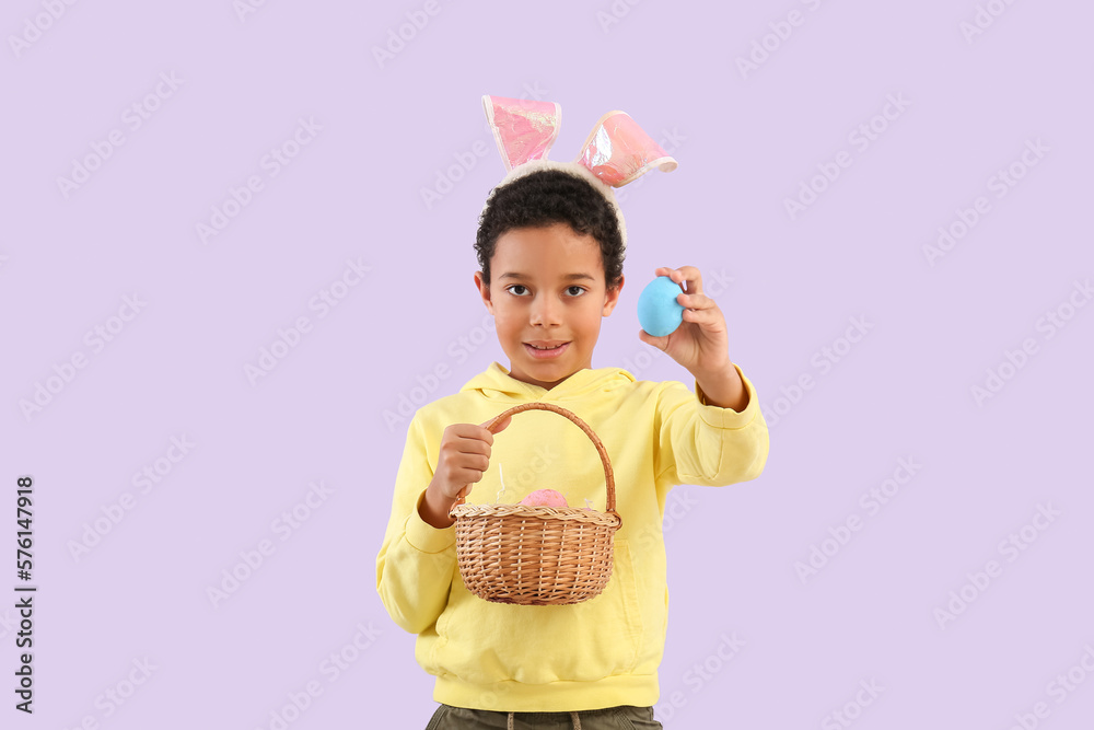 African-American little boy in bunny ears with basket of Easter eggs on lilac background African-American little boy in bunny ears with basket of Easter eggs on lilac background