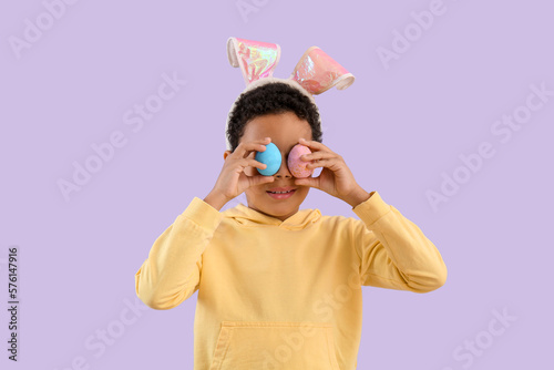 African-American little boy in bunny ears with Easter eggs on lilac background