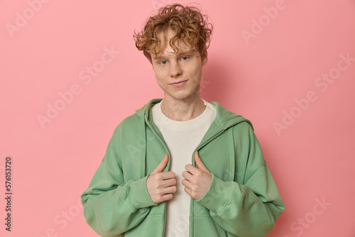 Waist up studio shot of a young curly red haired man looks and smiles at camera wears trendy clothes adjusts his green zipped hoodie over a pink background blank copy space for your advertising.