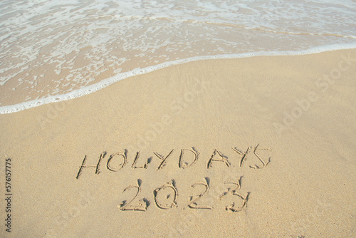Holidays 2023 lettering on the beach with wave and clear blue sea.