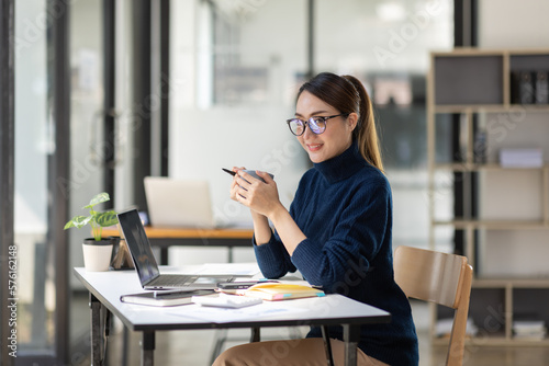 Young Asian professional business woman office worker analyst sitting at desk working on laptop thinking on project plan, analyzing marketing or financial data online, watching elearning webinar.