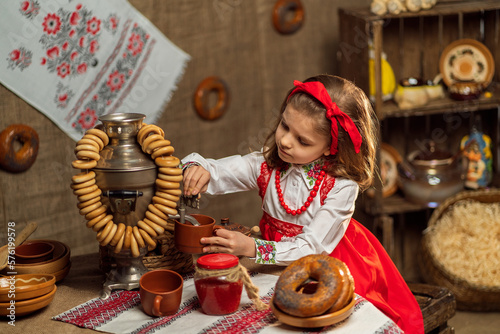 Little girl in folk dress pours tea from samovar while sitting in folk decor room. Shrovedite 