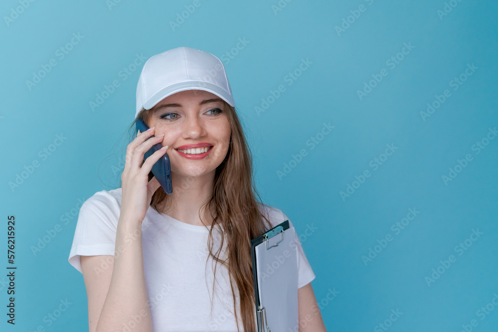 Cheerful young woman talking on phone holding clipboard conducting survey or advertising a product on a blue background in a white t-shirt and cap with space for a mockup Cheerful young woman talking on phone holding clipboard conducting survey or advertising a product on a blue background in a white t-shirt and cap with space for a mockup