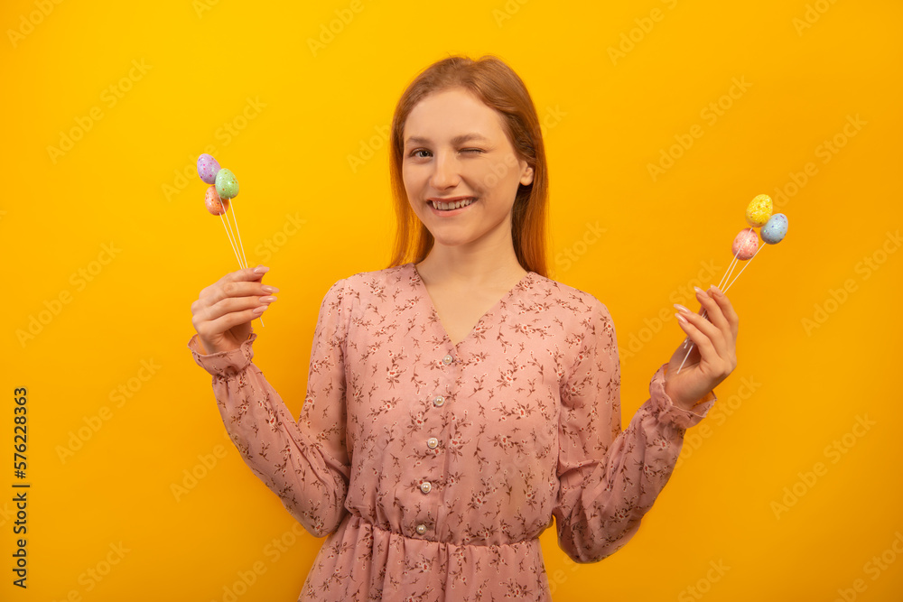 Happy smiling winking young woman in pink flower dress showing multicolored painted dyed easter eggs on sticks in two hands and looking at camera isolated on yellow bckground.
Easter day concept. Happy smiling winking young woman in pink flower dress showing multicolored painted dyed easter eggs on sticks in two hands and looking at camera isolated on yellow bckground.
Easter day concept.
