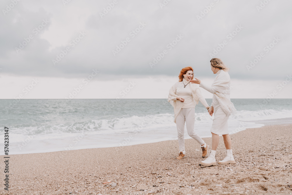 Women sea walk friendship spring. Two girlfriends, redhead and blonde, middle-aged walk along the sandy beach of the sea, dressed in white clothes. Against the backdrop of a cloudy sky and the winter Women sea walk friendship spring. Two girlfriends, redhead and blonde, middle-aged walk along the sandy beach of the sea, dressed in white clothes. Against the backdrop of a cloudy sky and the winter